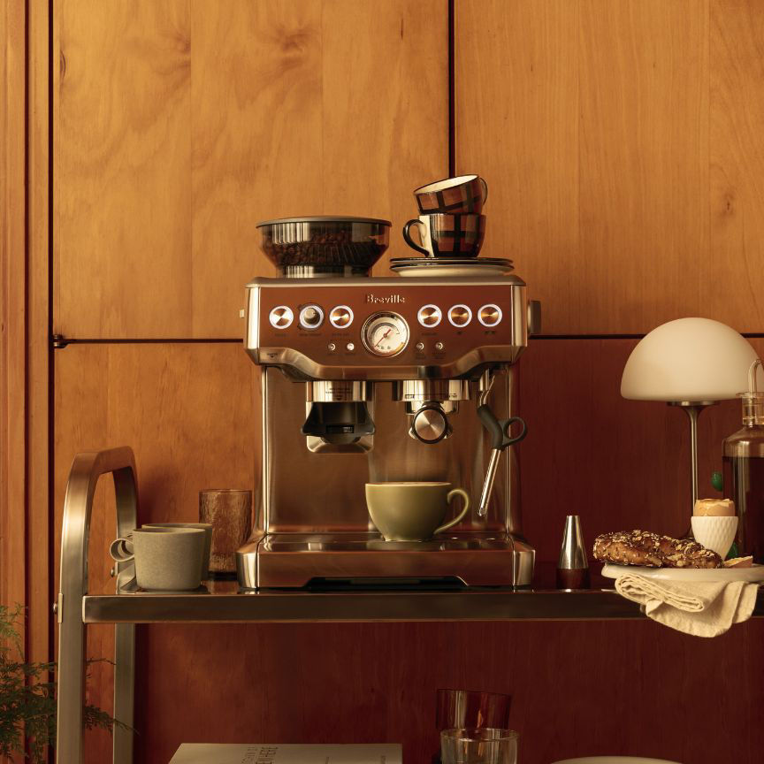 Breville Barista Express espresso machine on wooden sideboard, beans in hopper, espresso flowing into cup with cups and lamp arranged alongside